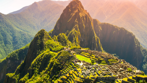 Estructuras de piedra de Machu Picchu se encuentran en una ladera montañosa, rodeadas de exuberante vegetación y montañas en el fondo. La luz del sol ilumina la escena, resaltando los tonos verdes de la naturaleza y el gris de las piedras.